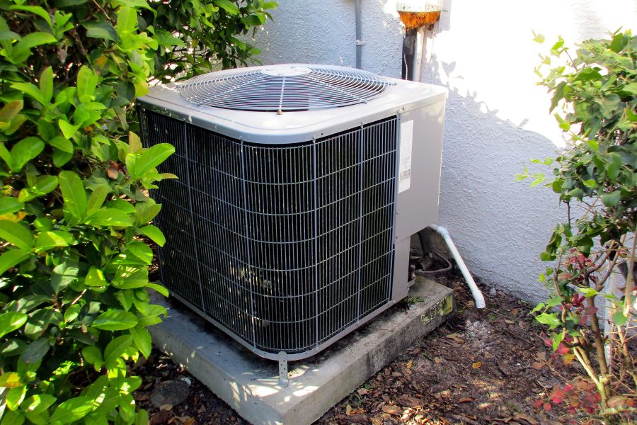 Outdoor air conditioning condenser unit on a concrete pad, surrounded by green shrubs and a white wall nearby.