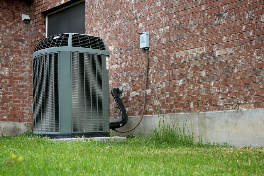 Outdoor central air conditioner condenser unit next to a brick wall with supply lines and a small grassy area in front.