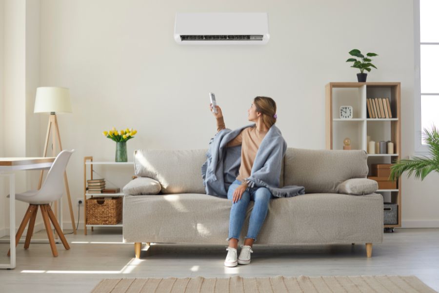 Woman seated on a beige sofa in a bright living room, holding a remote toward a wall-mounted white air conditioner above her.