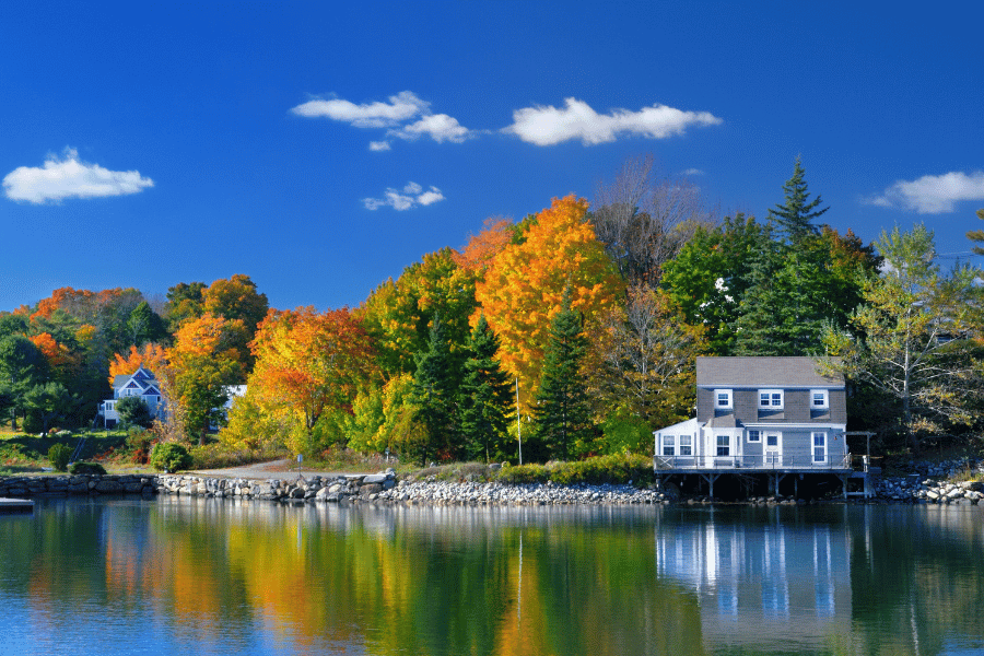 Serene lakeside scene with autumn trees in orange and yellow, a gray house on stilts by the water reflecting the sky and colorful foliage.