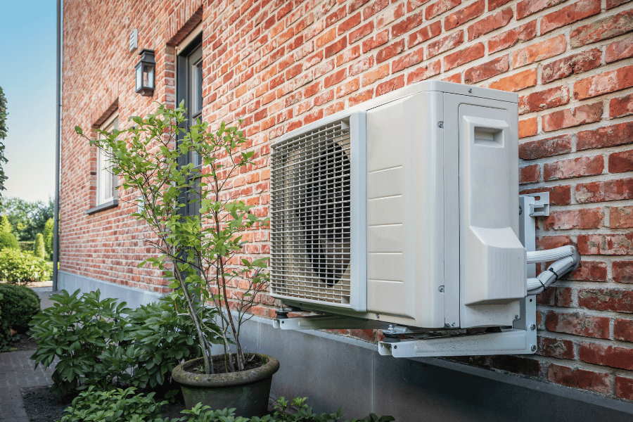 Outdoor air conditioner condenser unit mounted on a brick wall with a metal support bracket, nearby potted plant and shrubs in a sunny yard.