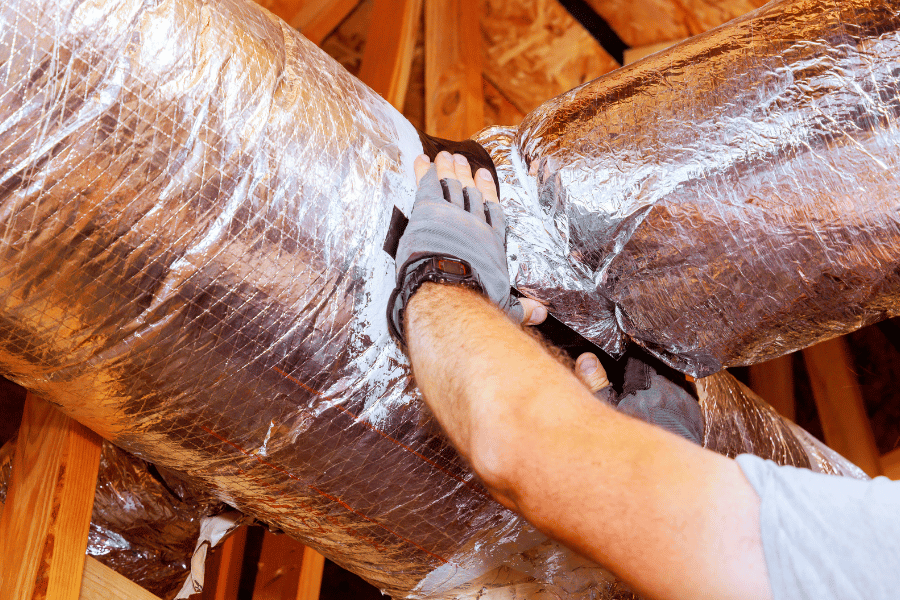 Person wearing gloves adjusting foil-insulated HVAC ductwork in an attic scene with wooden framing