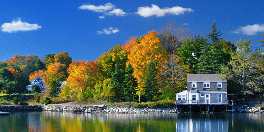 Serene lakeside scene with autumn trees in orange and yellow, a gray house on stilts by the water reflecting the sky and colorful foliage.
