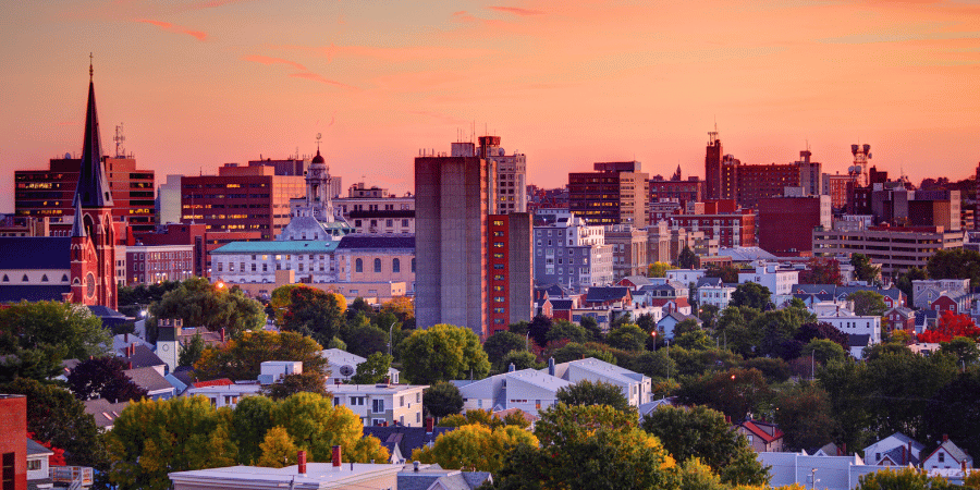 City skyline at sunset with orange-pink sky over a mix of modern and historic buildings and trees in the foreground.