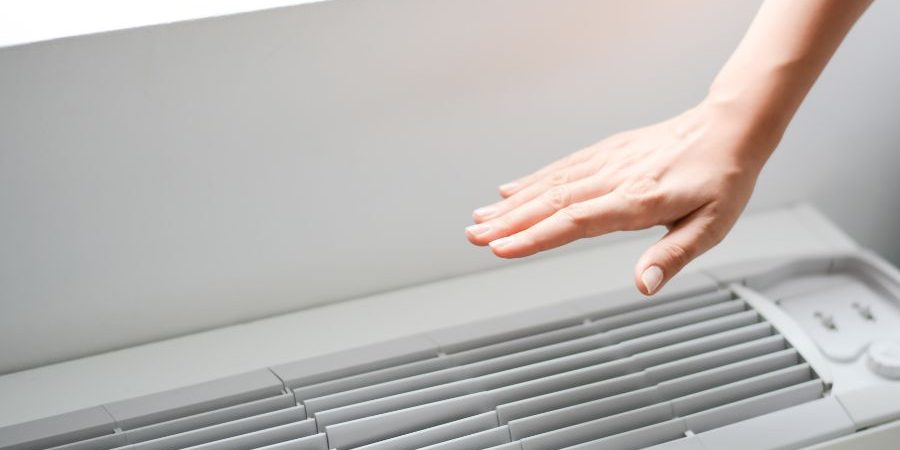 Hand hovering over the vents of a white window air conditioner, as if about to turn it on or adjust it.